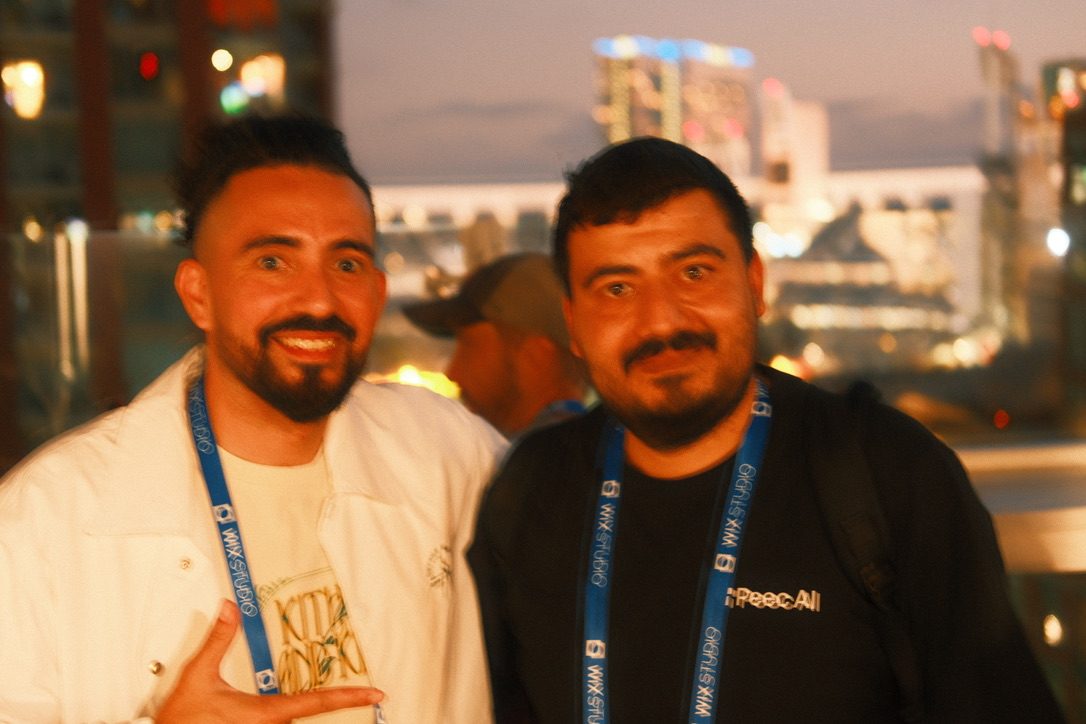 Two brightonSEO San Diego attendees wearing conference lanyards pose together at SERP & TURF, one in a Peec AI shirt, with the San Diego skyline at night behind them