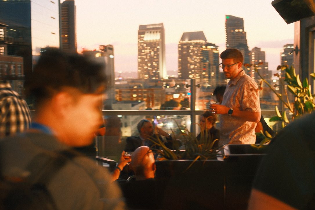 SERP & TURF guests with drinks on the rooftop at dusk, with the lit towers of downtown San Diego behind them