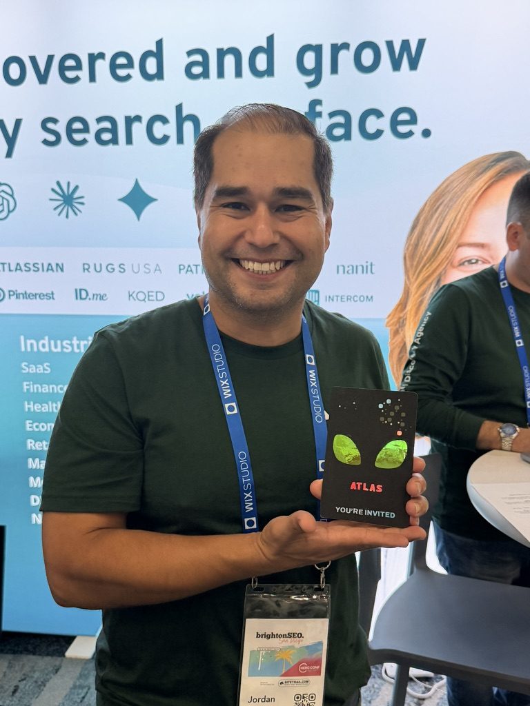 Jordan Koene, CEO of Previsible, holds a Momentic Atlas invitation card while wearing his brightonSEO San Diego 2026 badge