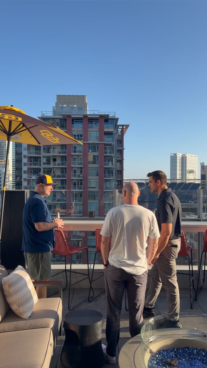 Guests talk on the rooftop at SERP & TURF under a San Diego Padres umbrella, with Petco Park and the downtown San Diego skyline behind them
